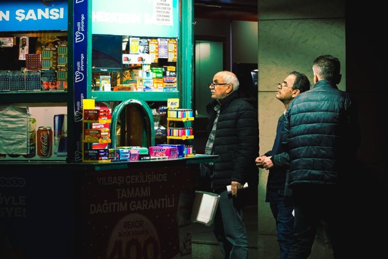 Three men at a lottery stand observe the variety of items for sale under bright lights.
