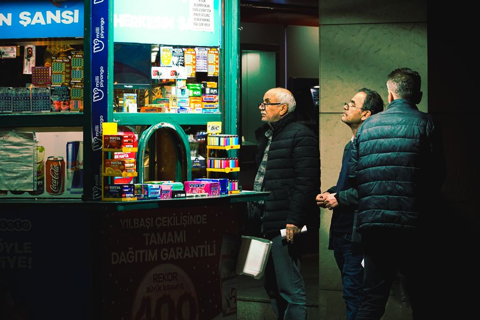 Three men at a lottery stand observe the variety of items for sale under bright lights.